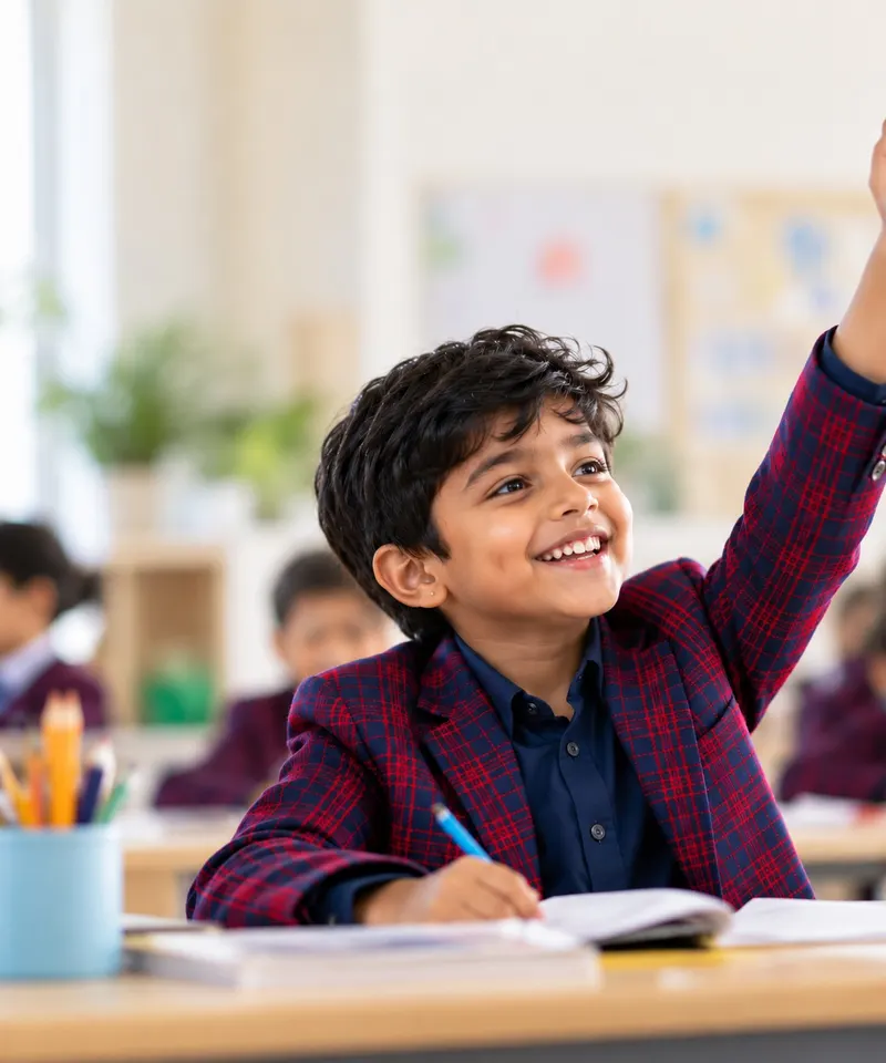 A smiling student in a Sloka blazer raising his hand in class.